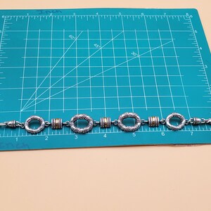 Op de afbeelding: Zilverkleurige armband met goudkleurige accenten. De armband heeft een reeks cirkelvormige en cilindrische schakels. De armband is ongeveer 17,78 cm lang.