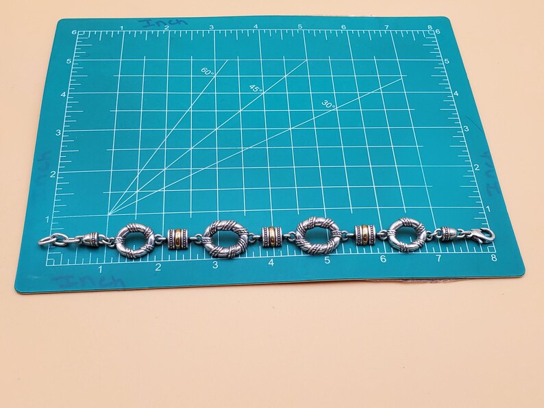 Op de afbeelding: Zilverkleurige armband met goudkleurige accenten. De armband heeft een reeks cirkelvormige en cilindrische schakels. De armband is ongeveer 17,78 cm lang.