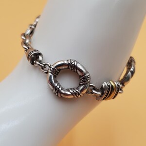 Op de afbeelding: Zilverkleurige armband met een cirkelvormige sluiting en decoratieve schakels. De armband heeft een textuurontwerp rond de sluiting en accenten van goudkleurig metaal. De armband wordt getoond op een witte ondergrond tegen een lichtoranje achtergrond.