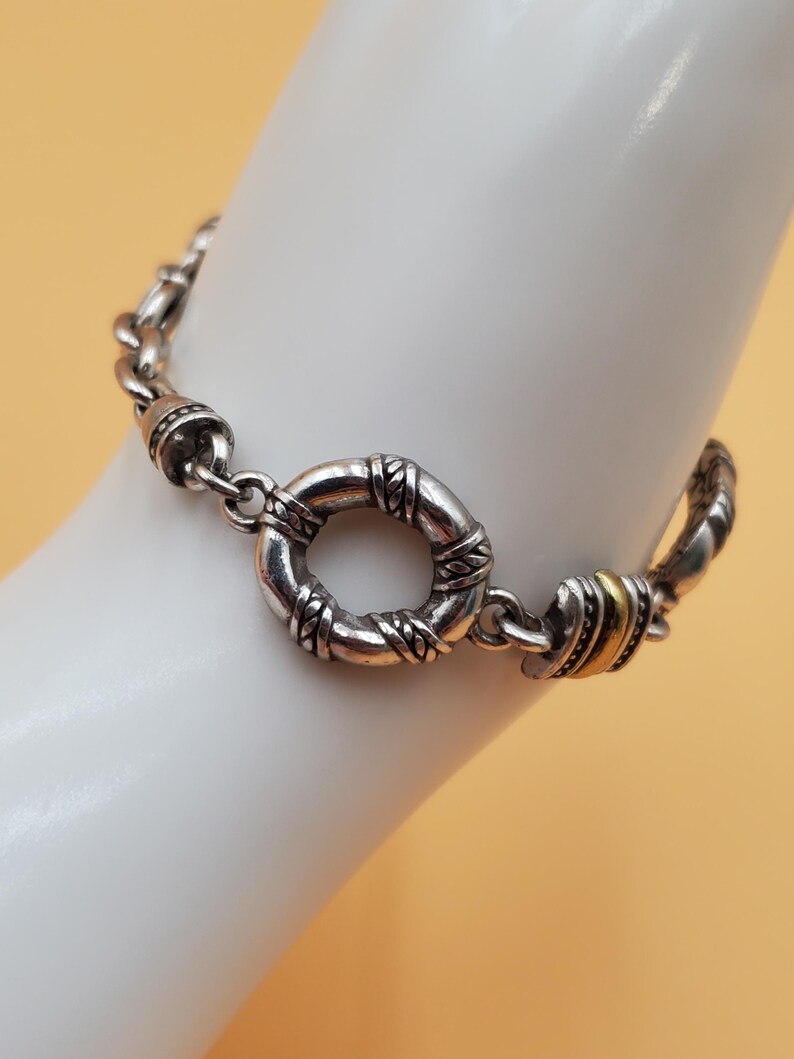 Op de afbeelding: Zilverkleurige armband met een cirkelvormige sluiting en decoratieve schakels. De armband heeft een textuurontwerp rond de sluiting en accenten van goudkleurig metaal. De armband wordt getoond op een witte ondergrond tegen een lichtoranje achtergrond.