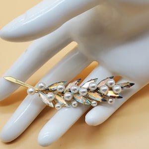 May include: A gold-toned brooch in a leaf design, adorned with iridescent crystals and faux pearls. The brooch is shaped like a slender branch, with the pearls clustered along the stem. The crystals reflect multiple colors.