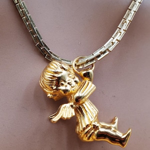 Pu&ograve; includere: Una collana color oro con un ciondolo a forma di cherubino. Il ciondolo raffigura un cherubino seduto con ali, lineamenti del viso dettagliati e abiti. La catena &egrave; a maglie piatte.