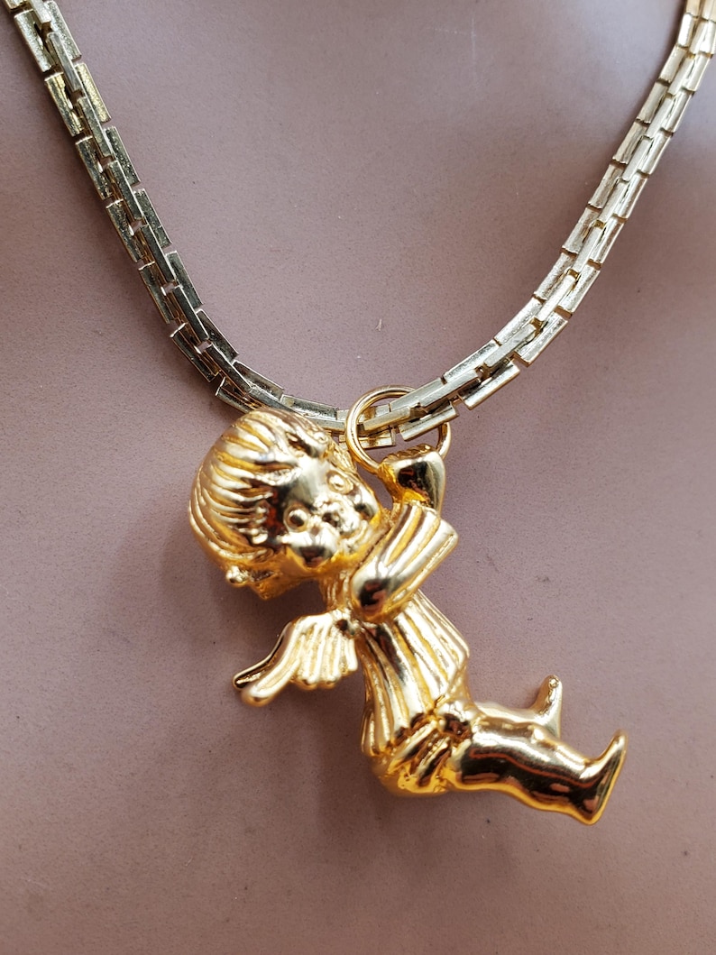 Pu&ograve; includere: Una collana color oro con un ciondolo a forma di cherubino. Il ciondolo raffigura un cherubino seduto con ali, lineamenti del viso dettagliati e abiti. La catena &egrave; a maglie piatte.