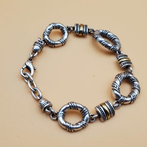 Op de afbeelding: Zilverkleurige armband met getextureerde, cirkelvormige schakels en goudkleurige accenten. De armband heeft een karabijnsluiting en een ketting voor verstelbaarheid. Het ontwerp is gedetailleerd, met een vintage uitstraling. De schakels zijn verbonden.