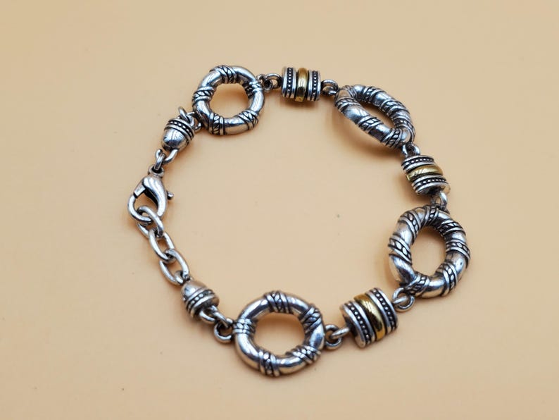 Op de afbeelding: Zilverkleurige armband met getextureerde, cirkelvormige schakels en goudkleurige accenten. De armband heeft een karabijnsluiting en een ketting voor verstelbaarheid. Het ontwerp is gedetailleerd, met een vintage uitstraling. De schakels zijn verbonden.