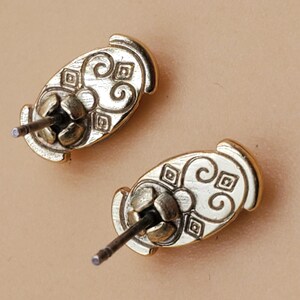May include: Two gold-colored stud earrings. Each earring has an oval shape with an ornate, swirling design and a post for insertion. The earrings are displayed against a neutral background.