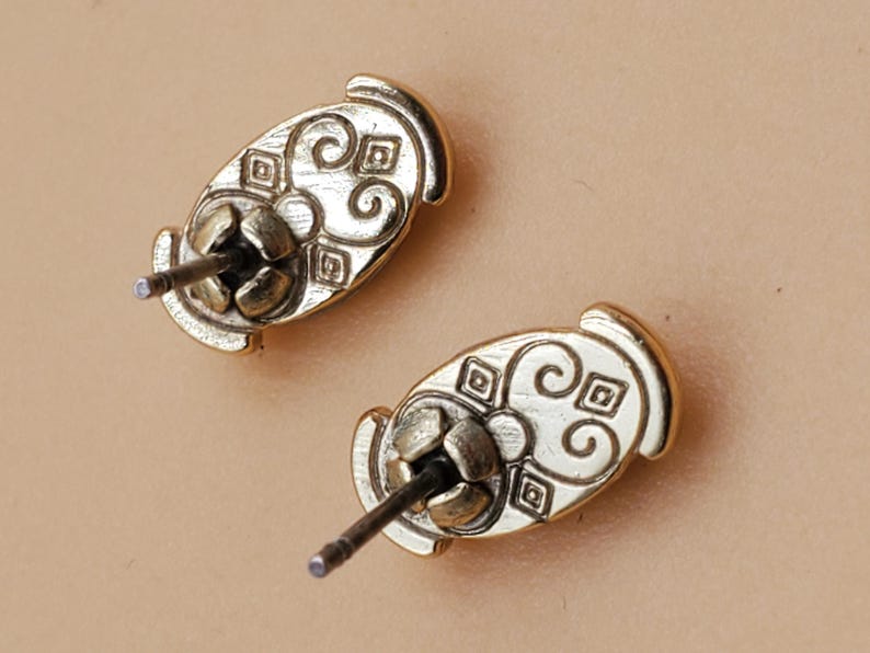 May include: Two gold-colored stud earrings. Each earring has an oval shape with an ornate, swirling design and a post for insertion. The earrings are displayed against a neutral background.