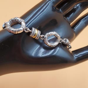 Op de afbeelding: Zilverkleurige armband met ovale en cirkelvormige schakels, met een textuurpatroon. De armband heeft een goudkleurig accent. Het sieraad wordt gepresenteerd op een zwart oppervlak tegen een neutrale achtergrond.