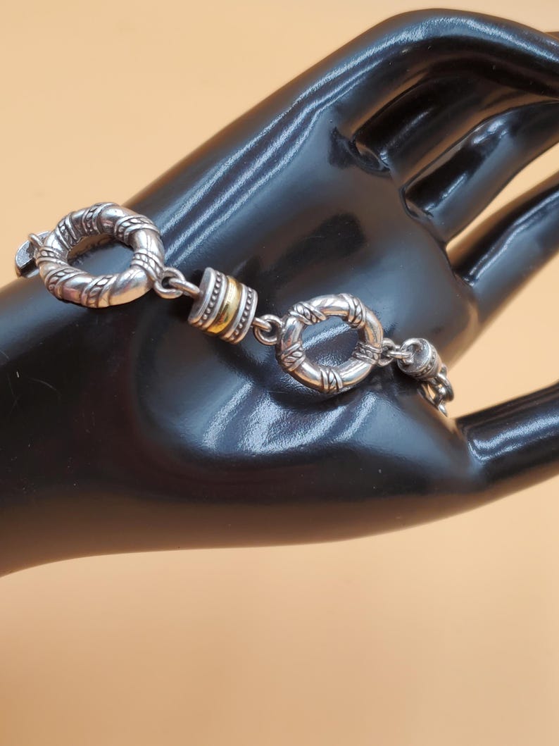 Op de afbeelding: Zilverkleurige armband met ovale en cirkelvormige schakels, met een textuurpatroon. De armband heeft een goudkleurig accent. Het sieraad wordt gepresenteerd op een zwart oppervlak tegen een neutrale achtergrond.