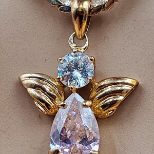 May include: A gold angel pendant with a pear-shaped, clear gemstone body and a round, light blue gemstone head. The pendant features gold wings and hangs from a gold and silver chain. The jewellery is displayed against a neutral background.