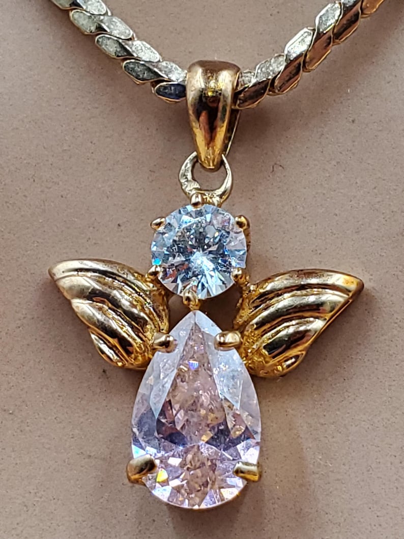 May include: A gold angel pendant with a pear-shaped, clear gemstone body and a round, light blue gemstone head. The pendant features gold wings and hangs from a gold and silver chain. The jewellery is displayed against a neutral background.