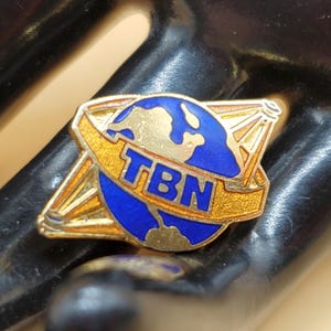 Peut inclure: Une épingle dorée avec un globe bleu et les lettres "TBN" sur une bannière jaune. Le globe a une masse terrestre dorée et un océan bleu.