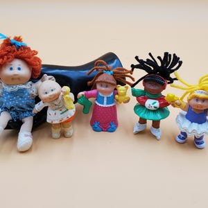 Puede incluir: Cinco muñecas vintage de Cabbage Patch Kids. Las muñecas tienen diferentes colores de cabello y atuendos, incluyendo un vestido azul, un vestido blanco y naranja, un vestido rosa, una falda verde y un tutú azul y blanco. Cada muñeca sostiene un pequeño juguete.