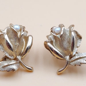 Puede incluir: Un par de pendientes de rosa dorados. Cada pendiente presenta un diseño de rosa detallado con una pequeña perla redonda en el centro. Los pendientes tienen un tallo y detalles de hojas, creando un accesorio floral clásico.
