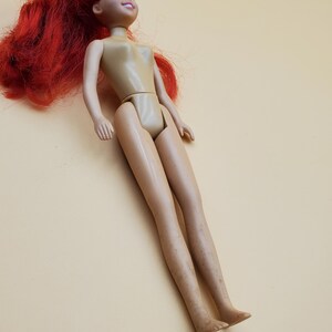 May include: A plastic doll with long red hair and blue eyes. The doll is wearing a nude-colored swimsuit.