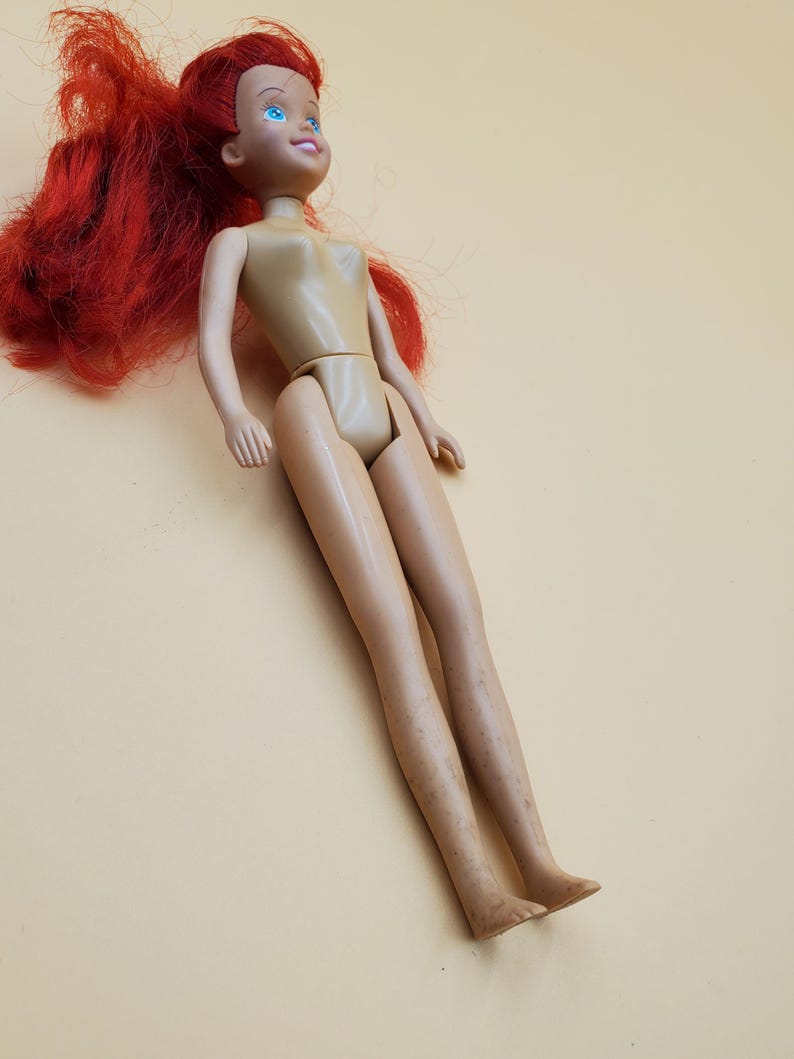 May include: A plastic doll with long red hair and blue eyes. The doll is wearing a nude-colored swimsuit.