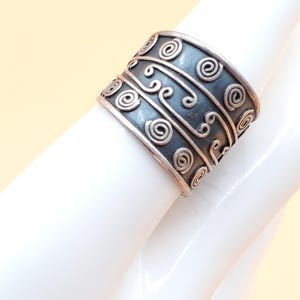 May include: Wide silver ring with an oxidized finish, featuring an intricate design of swirling patterns and spirals. The ring has a bold, cuff-like appearance, showcasing detailed craftsmanship and a vintage aesthetic.
