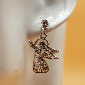 K&ouml;nnte beinhalten: Silberfarbene Engels-Ohrringe mit klaren Kristallakzenten. Die Ohrringe haben ein h&auml;ngendes Engel-Design mit filigranen Details und einem kleinen Kristall oben. Der Engel hat ausgestreckte Arme und Fl&uuml;gel und ist an einem Pfosten mit einem Kristall befestigt.