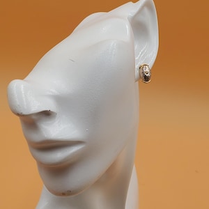 May include: A gold-toned earring with a small, oval design featuring white and clear accents. The earring is clipped onto a white mannequin head against an orange background. The earring has a small clasp.
