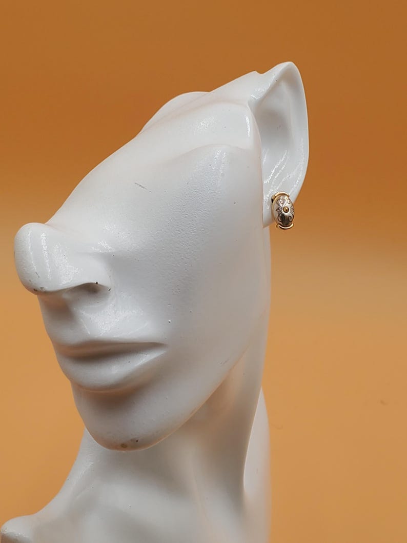 May include: A gold-toned earring with a small, oval design featuring white and clear accents. The earring is clipped onto a white mannequin head against an orange background. The earring has a small clasp.