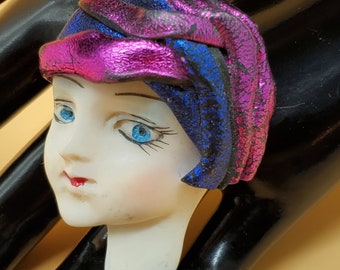 Puede incluir: Una cabeza de muñeca de porcelana con ojos azules y labios rojos, que lleva un sombrero de tela rosa y azul con textura brillante.