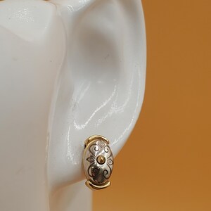 May include: A gold and silver-toned oval earring with a decorative pattern. The earring is a clip-on style, with a gold-colored band at the top and bottom. The earring is displayed on a white ear-shaped form against an orange background.