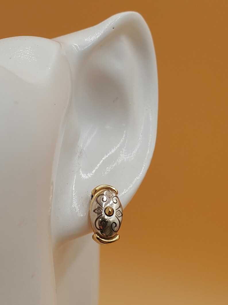 May include: A gold and silver-toned oval earring with a decorative pattern. The earring is a clip-on style, with a gold-colored band at the top and bottom. The earring is displayed on a white ear-shaped form against an orange background.