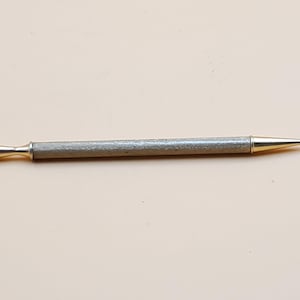 May include: A silver and gold pen with a textured, cylindrical body. The pen has a gold tip and a gold accent at the opposite end. The pen is lying on a light beige surface.