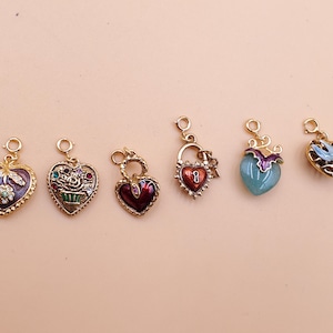 May include: Six heart-shaped charms with gold-coloured metalwork. The charms feature various designs, including a butterfly, flowers, a lock, and a bird. Each charm has a loop for attaching to a necklace or bracelet. The charms are in shades of red, blue, purple, and green.