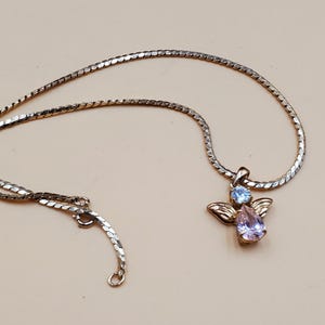 May include: Silver-coloured necklace with an angel pendant. The pendant features a light blue and a pear-shaped lavender gemstone, with gold-coloured wings. The necklace has a delicate chain and a lobster clasp.