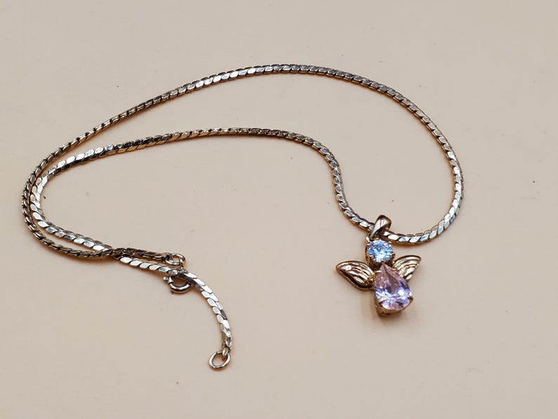 May include: Silver-coloured necklace with an angel pendant. The pendant features a light blue and a pear-shaped lavender gemstone, with gold-coloured wings. The necklace has a delicate chain and a lobster clasp.