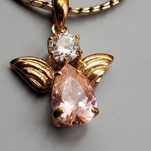 May include: Gold angel pendant necklace. The pendant has a pear-shaped pink gemstone body, a clear round gemstone head, and gold wings. The necklace chain is a gold snake chain. The background is a solid grey.