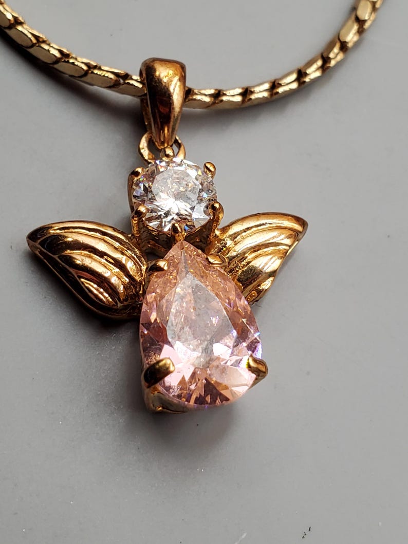 May include: Gold angel pendant necklace. The pendant has a pear-shaped pink gemstone body, a clear round gemstone head, and gold wings. The necklace chain is a gold snake chain. The background is a solid grey.