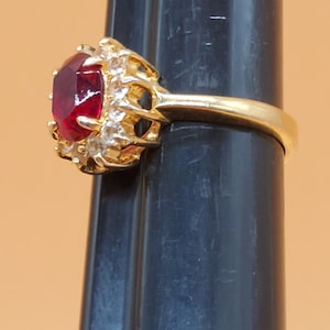 May include: A gold ring featuring a large red gemstone surrounded by smaller clear gemstones.