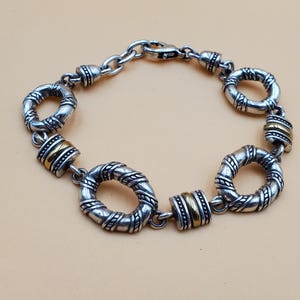 Op de afbeelding: Zilverkleurige armband met getextureerde, cirkelvormige schakels en goudkleurige cilindrische accenten. De armband heeft een karabijnsluiting en wordt gepresenteerd tegen een neutrale achtergrond. De lengte is ongeveer 19 cm.