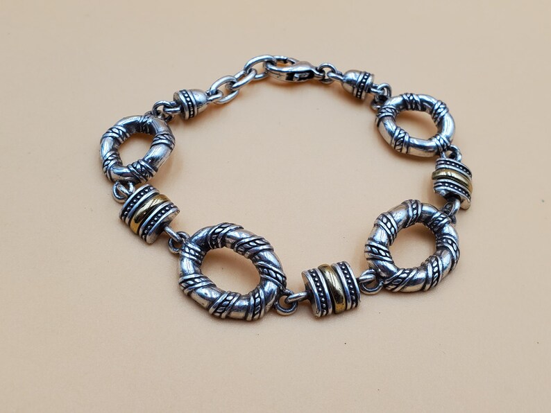 Op de afbeelding: Zilverkleurige armband met getextureerde, cirkelvormige schakels en goudkleurige cilindrische accenten. De armband heeft een karabijnsluiting en wordt gepresenteerd tegen een neutrale achtergrond. De lengte is ongeveer 19 cm.