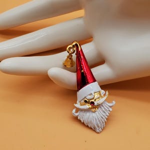 May include: A gold-tone metal brooch shaped like a Santa Claus head with a red enamel hat, a white enamel beard and moustache, and a small dangling gold bell attached to the top of the hat. The Santa figure features a small red rhinestone nose.