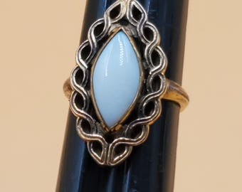 May include: A gold-toned ring featuring a light blue, oval-shaped stone. The stone is set within a decorative, openwork frame. The ring is displayed on a black stand against a neutral background. The ring's design is vintage.