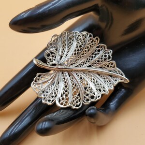 Puede incluir: Un broche plateado en forma de hoja con intrincados detalles calados. El broche tiene un diseño delicado de filigrana, con un tallo y un patrón de hoja veteada. La joya se exhibe sobre una superficie negra.
