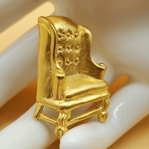 May include: A miniature gold-colored armchair with a high back and ornate details. The chair features a detailed backrest with star-shaped embellishments and curved legs. The overall design is reminiscent of a classic wingback chair.