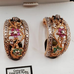 May include: A pair of gold-toned hoop earrings with intricate filigree and gemstone accents. The earrings feature a floral design with pink, green, and clear gemstones. The text on the box reads "Genuine Emerald, Ruby, Sapphire & CZ."