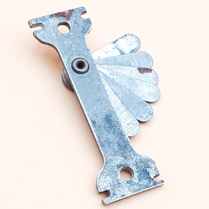 May include: A vintage metal feeler gauge with multiple blades fanned out. The tool has a central pivot and two slotted ends. The metal shows signs of age with some rust. The background is a light beige.