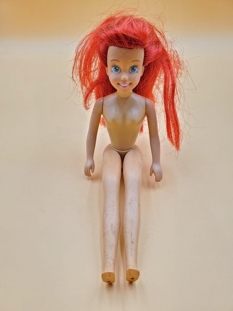 May include: A red-haired doll with blue eyes and a smiling face. The doll is wearing a nude-colored outfit and is sitting with its legs crossed.