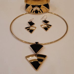 May include: A gold-tone jewelry set with black and white geometric designs. The set includes a choker necklace with a pendant, a pair of earrings, and a bangle bracelet.