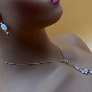 May include: A delicate gold chain necklace with a white oval pendant and a cluster of smaller white oval beads. The necklace is paired with matching earrings featuring a single white oval bead.