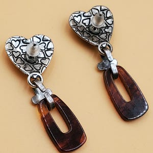 May include: A pair of drop earrings. Each earring has a heart-shaped top with a pattern of small hearts and a brown, rectangular pendant. The earrings are connected by a silver-coloured metal piece. The background is a solid, light tan colour.