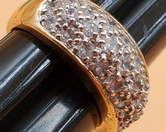 May include: A gold-toned ring with a band encrusted with numerous small, clear gemstones. The ring is set against a black background, with a close-up view highlighting the details of the jewelry.
