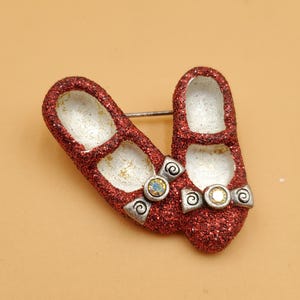 May include: A red glittery shoe brooch featuring two shoes. Each shoe has a white interior and a silver bow with a small, iridescent gem. The brooch is a whimsical accessory, perfect for adding a touch of sparkle to any outfit.