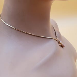 May include: A gold-coloured necklace featuring a fine chain and a small pendant. The pendant is a small, decorative figure with a clear stone. The necklace is displayed on a mannequin.