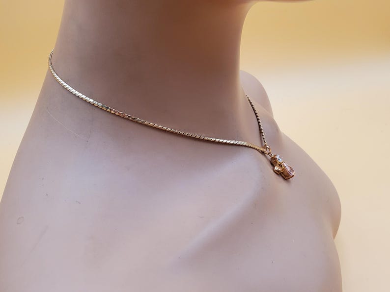 May include: A gold-coloured necklace featuring a fine chain and a small pendant. The pendant is a small, decorative figure with a clear stone. The necklace is displayed on a mannequin.